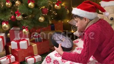 年轻的男孩在等圣诞老人，看着时钟，躺在室内装饰的圣诞树附近，灯光，打扮成圣诞老人的帮手-